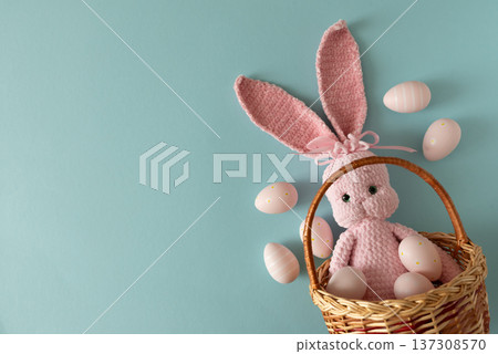 Woven basket with pink crocheted bunny doll surrounded by pastel Easter eggs against soft blue background Woven basket with pink crocheted bunny doll surrounded by pastel Easter eggs against soft blue background 137308570