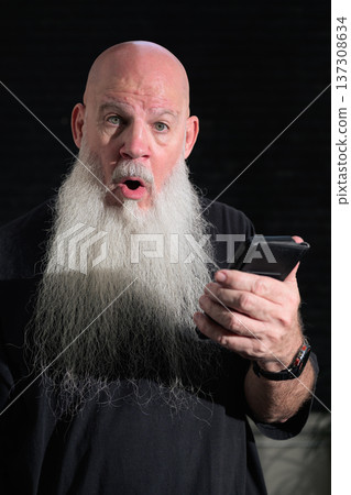 Portrait of a bald man with a long white beard using phone against a dark background Portrait of a bald man with a long white beard using phone against a dark background 137308634