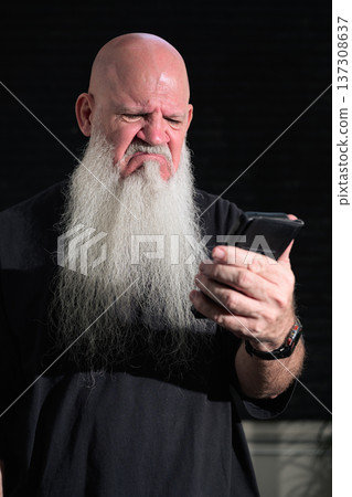 Portrait of a bald man with a long white beard using phone against a dark background Portrait of a bald man with a long white beard using phone against a dark background 137308637