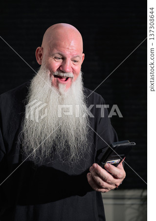 Portrait of a happy bald man with a long white beard using phone against a dark background Portrait of a happy bald man with a long white beard using phone against a dark background 137308644