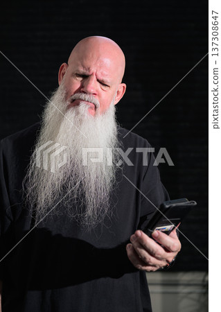 Portrait of a sad bald man with a long white beard using phone against a dark background Portrait of a sad bald man with a long white beard using phone against a dark background 137308647