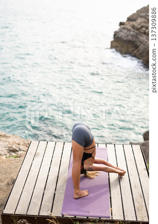 Woman performing standing forward bend on a yoga mat outdoors near the sea. Focusing on fitness, wellness, and mindfulness Woman performing standing forward bend on a yoga mat outdoors near the sea. Focusing on fitness, wellness, and mindfulness 137309886
