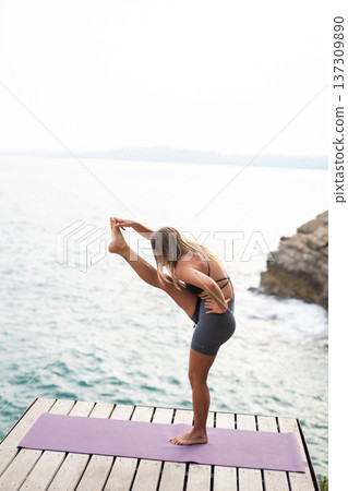 Woman performing a standing balance yoga pose on a mat by the ocean, focusing on flexibility, wellness, and exercise 137309890