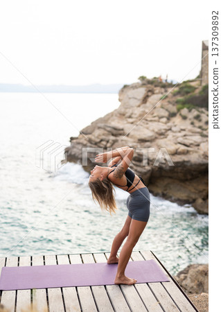 Young woman bending backward in ustrasana on a yoga mat, finding balance and relaxation by the ocean Young woman bending backward in ustrasana on a yoga mat, finding balance and relaxation by the ocean 137309892