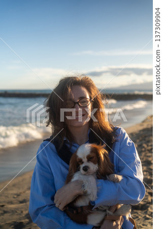 A young woman hugs her Cavalier King Charles Spaniel on the beach at sunset. Warm light, waves, and smiles capture a peaceful, joyful moment of connection and relaxation. 137309904