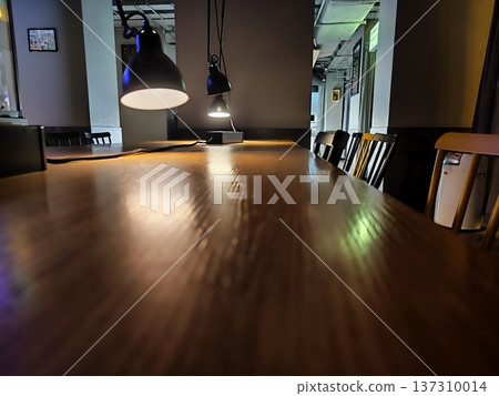 Dimly lit interior featuring a long wooden table with a smooth surface. Several black chairs line the sides, and overhead, black pendant lights Dimly lit interior featuring a long wooden table with a smooth surface. Several black chairs line the sides, and overhead, black pendant lights 137310014