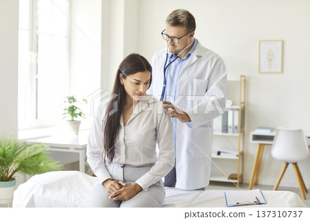 Serious doctor checking up female patient in clinic holding stethoscope during medical exam Serious doctor checking up female patient in clinic holding stethoscope during medical exam 137310737