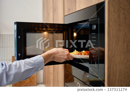 Hand holds plate with cooked food at open built in microwave oven in kitchen cabinet Hand holds plate with cooked food at open built in microwave oven in kitchen cabinet 137311274