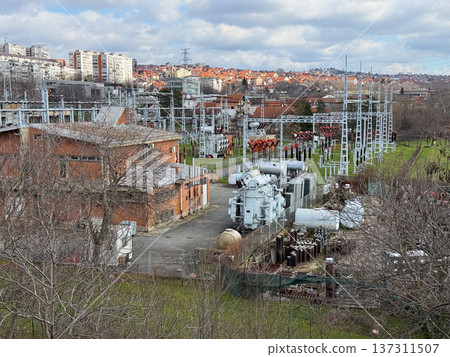Large electrical power substation with transformers cables and metal structures surrounded by urban neighborhood and cloudy sky landscape. Energy infrastructure, electricity distribution system 137311507