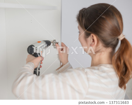 A Caucasian woman uses a screwdriver to tighten a screw while assembling a cabinet. A Caucasian woman uses a screwdriver to tighten a screw while assembling a cabinet. 137311683