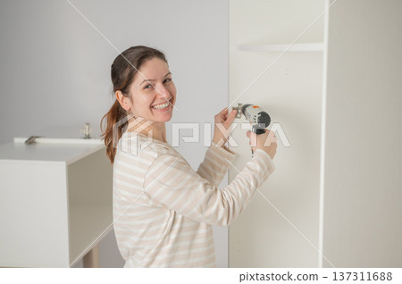 A Caucasian woman uses a screwdriver to tighten a screw while assembling a cabinet. 137311688