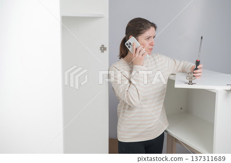 A Caucasian woman talks on the phone while assembling furniture. 137311689