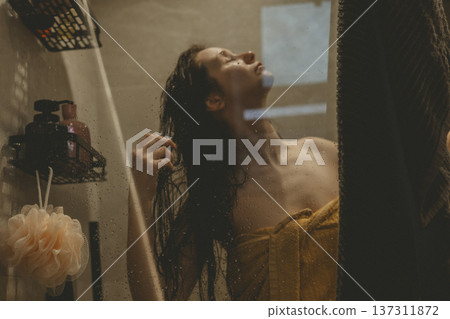Young woman practicing self-care ritual in cozy bathroom, washing hair behind glass shower door with water drops, cinematic warm lighting, mental health and morning routine concept authentic lifestyle 137311872