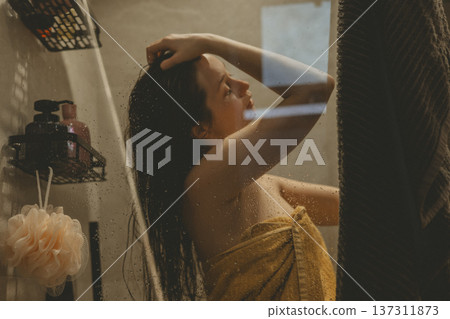 Young woman practicing self-care ritual in cozy bathroom, washing hair behind glass shower door with water drops, cinematic warm lighting, mental health and morning routine concept authentic lifestyle 137311873