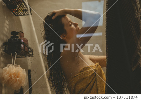 Young woman practicing self-care ritual in cozy bathroom, washing hair behind glass shower door with water drops, cinematic warm lighting, mental health and morning routine concept authentic lifestyle 137311874