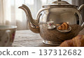 A silver teapot sits on a wooden table reflecting a cup and a croissant. Natural light fills the room showing a cozy breakfast setting. 137312682
