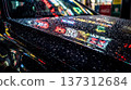 Rain drops cover a black car surface, showing bright reflections of city signs and lights at night. The scene captures the busy atmosphere of urban life. 137312684