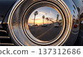 A classic car sits on a coastal road as the sun sets, casting a warm glow. Palm trees frame the street and vintage cars drive past during this evening scene. 137312686