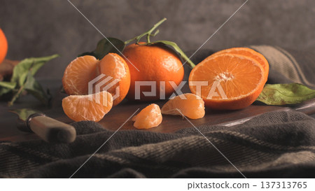 Tangerines and orange slices arranged on a dark wooden board with a knife and plaid fabric, capturing a cozy, rustic still life 137313765
