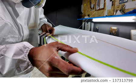 Close up of a surfboard shaper in a white protective suit applying lime green masking tape to a foam board blank Close up of a surfboard shaper in a white protective suit applying lime green masking tape to a foam board blank 137313799