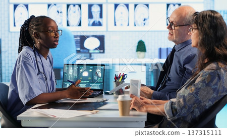 African american medical staff explaining test results to old couple, reviewing diagnostic results in medical cabinet. Uses advanced healthcare technology to evaluate MRI scans. Camera B. African american medical staff explaining test results to old couple, reviewing diagnostic results in medical cabinet. Uses advanced healthcare technology to evaluate MRI scans. Camera B. 137315111