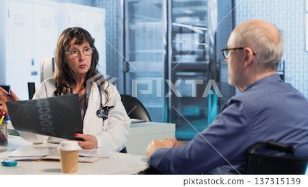 Woman medic presenting diagnostic report to wheelchair user during consultation, discussing illness and treatment plan. Physician doing scan interpretation under insurance. Impairment. Camera B. 137315139