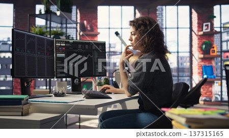 Female day trader examines stock market graphs on dual monitors, tracking financial growth to share prices. Trading interface provides real time data for buying and selling shares. Camera A. 137315165