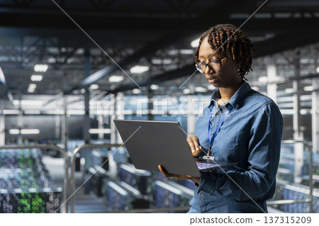Data center black woman checking lines of code on laptop while she stands on industrial platform, overseeing AI tasks for automation. Server room IT expert visualizing deep learning process. Data center black woman checking lines of code on laptop while she stands on industrial platform, overseeing AI tasks for automation. Server room IT expert visualizing deep learning process. 137315209