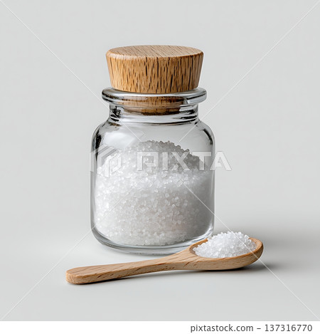 Glass jar with natural wooden lid filled with white salt next to a wooden spoon on gray background 137316770