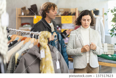 Woman choose jacket in store, exasperated husband accompanies his wife during shopping Woman choose jacket in store, exasperated husband accompanies his wife during shopping 137317623