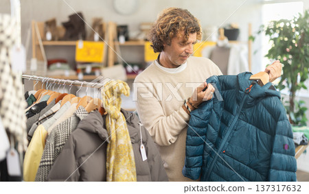 Focused male client carefully looking down jacket for autumn-winter season in clothing mall. Sale of winter clothing collection Focused male client carefully looking down jacket for autumn-winter season in clothing mall. Sale of winter clothing collection 137317632