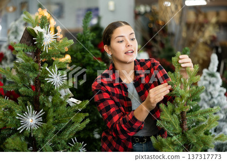 Young female in plaid shirt buying artificial Christmas trees before holidays in a store 137317773