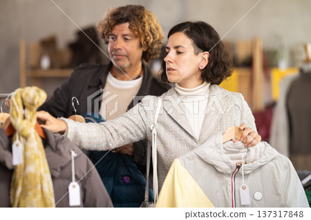European female buyer selecting new jacket or coat for cold season and man helping choose right size in boutique 137317848