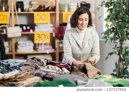 European female buyer standing in clothing market and carefully examines selection of warm sweaters for winter season European female buyer standing in clothing market and carefully examines selection of warm sweaters for winter season 137317958