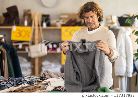 man stands near the counter with clothes and chooses a sweater man stands near the counter with clothes and chooses a sweater 137317982