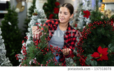 Female seller decorating an artificial Christmas tree with red holly berries at Christmas market Female seller decorating an artificial Christmas tree with red holly berries at Christmas market 137318078
