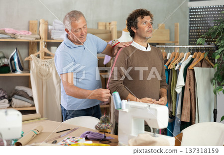 Elderly man tailor takes body measurements from adult man 137318159