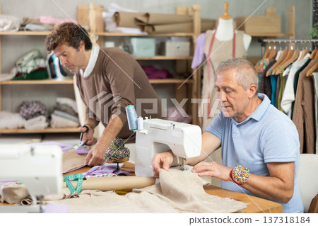 Mature tailor sewing clothes next to fabric cutter 137318184