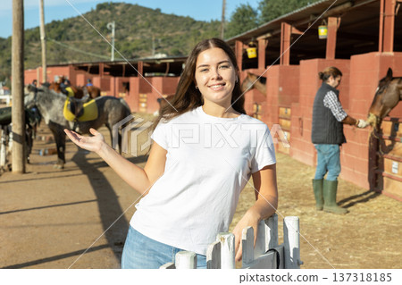 Portrait of cheerful European young woman wearing white T-shirt and posing on yard of horse club 137318185