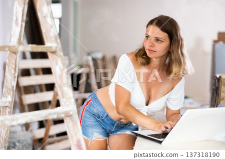 Young attractive girl checks the quality and correctness of the completed repairs using a laptop inside cottage Young attractive girl checks the quality and correctness of the completed repairs using a laptop inside cottage 137318190
