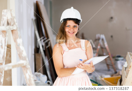 Girl in beige dress and protective helmet notes in document the completed stages of repair work in the cottage Girl in beige dress and protective helmet notes in document the completed stages of repair work in the cottage 137318287