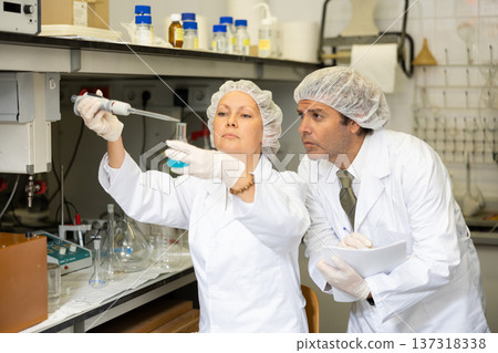 Middle-aged male and female researchers mixing reagents in test tube with lab pipette and writing report in laboratory 137318338