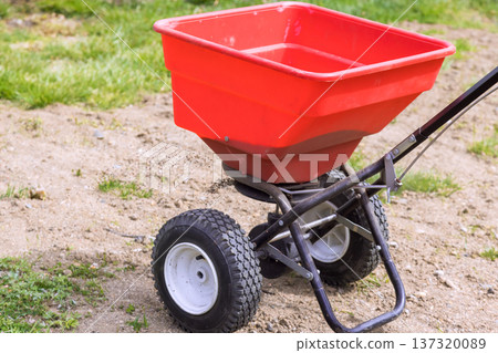 Seeds spreader is located on bare soil with grass around it, waiting for use in gardening tasks. 137320089