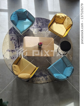 A top-down view of a modern lounge area featuring vibrant yellow and teal armchairs arranged around a wooden coffee table on a circular patterned rug, bathed in natural sunlight and soft shadows. 137320279