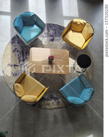 A top-down view of a modern lounge area featuring vibrant yellow and teal armchairs arranged around a wooden coffee table on a circular patterned rug, bathed in natural sunlight and soft shadows. A top-down view of a modern lounge area featuring vibrant yellow and teal armchairs arranged around a wooden coffee table on a circular patterned rug, bathed in natural sunlight and soft shadows. 137320280