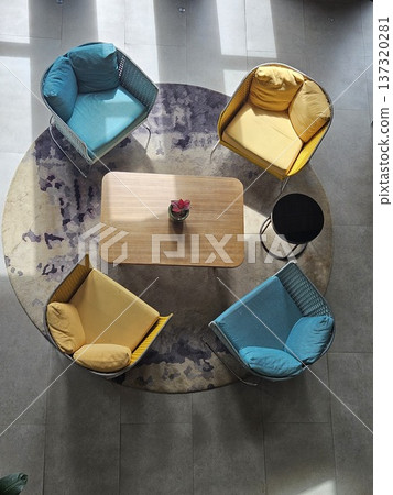 A top-down view of a modern lounge area featuring vibrant yellow and teal armchairs arranged around a wooden coffee table on a circular patterned rug, bathed in natural sunlight and soft shadows. 137320281