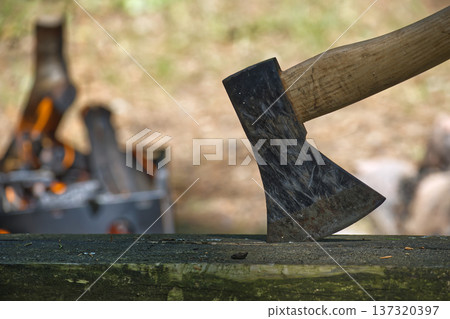 Close-up of an axe head resting on a wooden surface with a blurred campfire in the background Close-up of an axe head resting on a wooden surface with a blurred campfire in the background 137320397