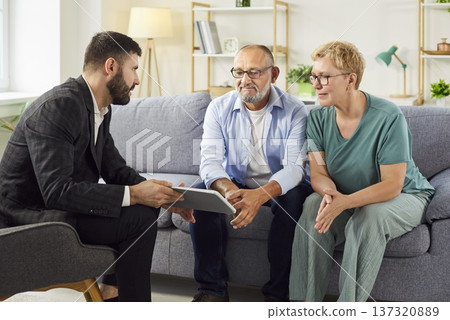 Real estate agent explaining terms to mature couple using tablet in living room Real estate agent explaining terms to mature couple using tablet in living room 137320889