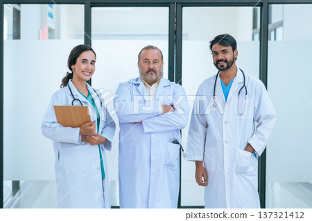Group portrait of diverse male and female doctors in white medical uniforms standing in hospital corridor smiling and looking at the camera. Group portrait of diverse male and female doctors in white medical uniforms standing in hospital corridor smiling and looking at the camera. 137321412