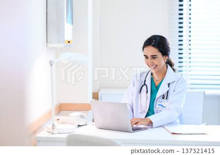 Smiling professional female doctor in white medical coat working with laptop computer, sitting at the table near the window in hospital office, looking at laptop screen. Smiling professional female doctor in white medical coat working with laptop computer, sitting at the table near the window in hospital office, looking at laptop screen. 137321415
